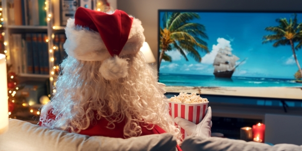 Santa sitting on a sofa watching a movie on a large screen and holding a bowl of popcorn