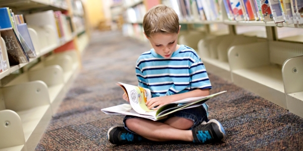 school-age child sitting on the floor in a library and reading a picture book