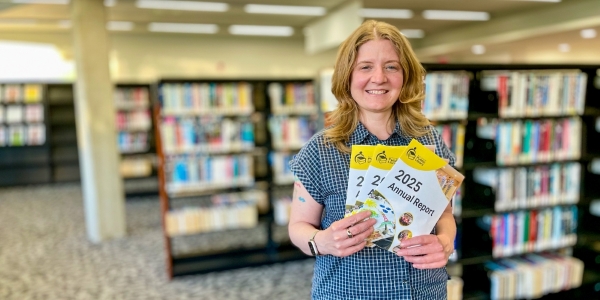 BPL chief executive officer, Lita Barrie holding copies of the 2025 Annual Report, standing by bookshelves