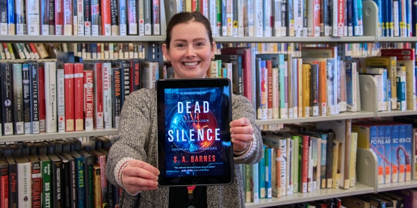 Sarah standing in front of library shelves and holding a copy of the book Dead Silence