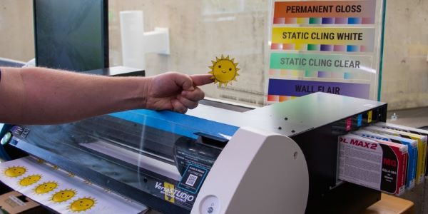 hand holding a smiling face decal with vinyl printer in background