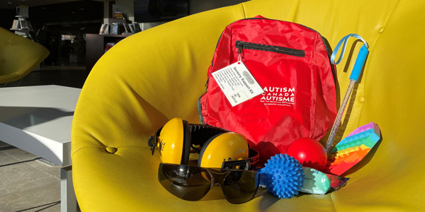 red backpack resting on a padded chair with items displayed beside it, including noise reducing ear covers, three fidget items, liquid motion bubble timer, finger spinner, glow baton, and tinted safety glasses.