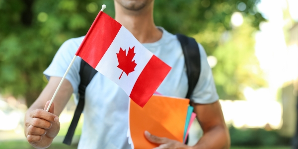 young adult wearing a white t-shirt and backpack and holding a small canada flag and three notebooks