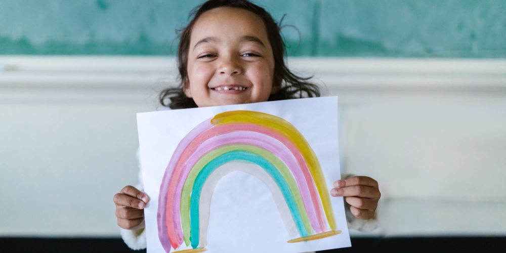 smiling child holding up a hand-drawn picture of a rainbow