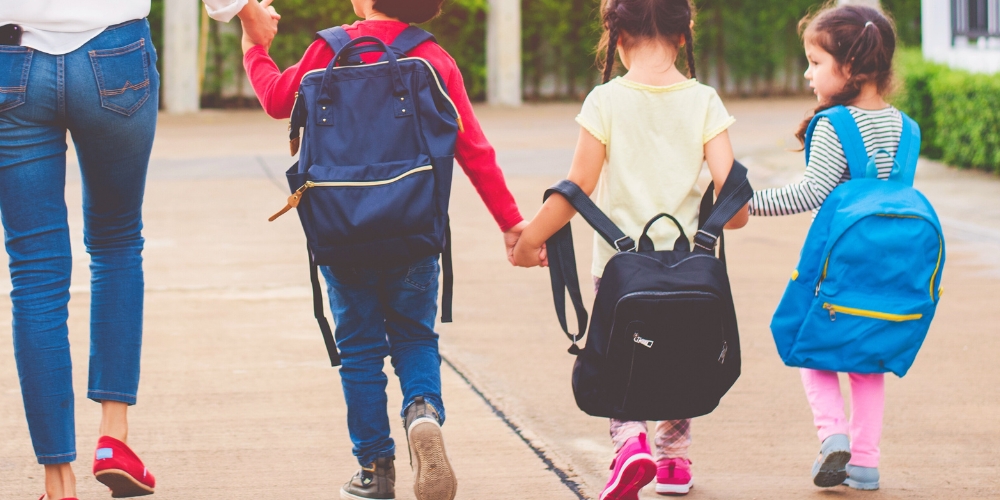 back view of three young children wearing backpacks and holding hands walking with a parent