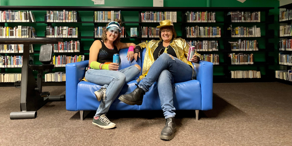 two people sitting on a blue couch with a beer in the library