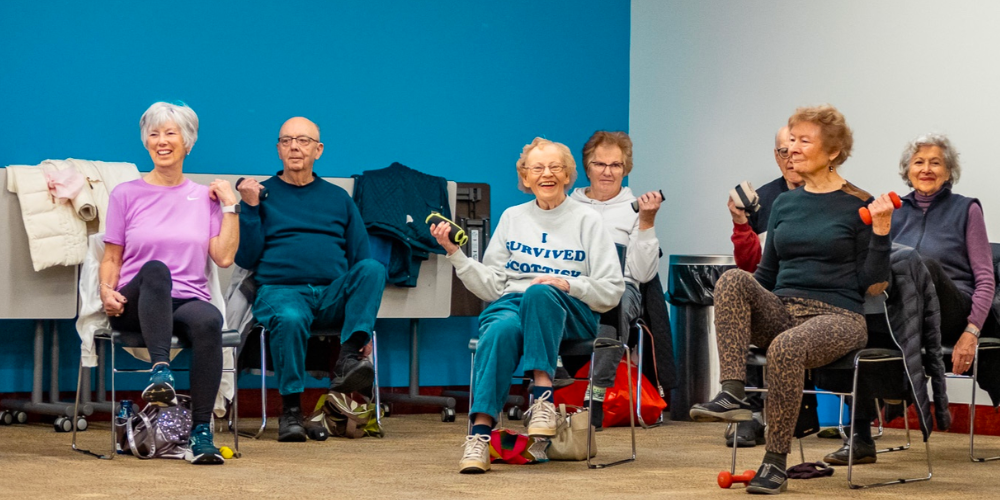 Seven older men and women sitting in chairs curling small hand weights
