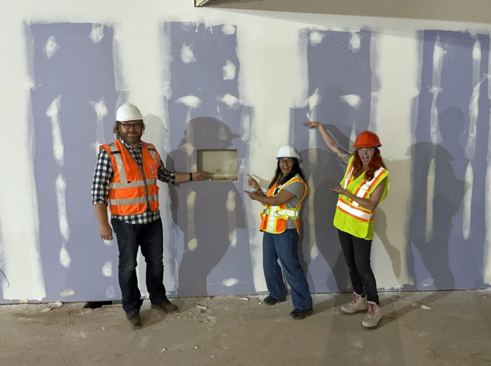 Three library staff members wearing safety vests and helmets pointing at a hole in the wall