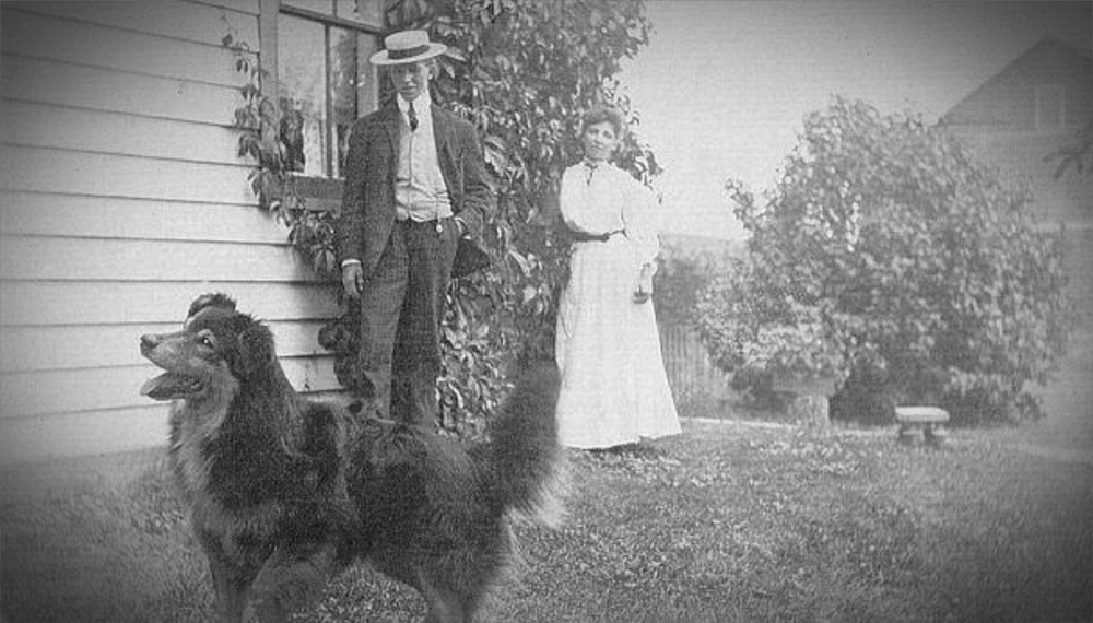 man, woman, and dog standing near a house in early 1900s
