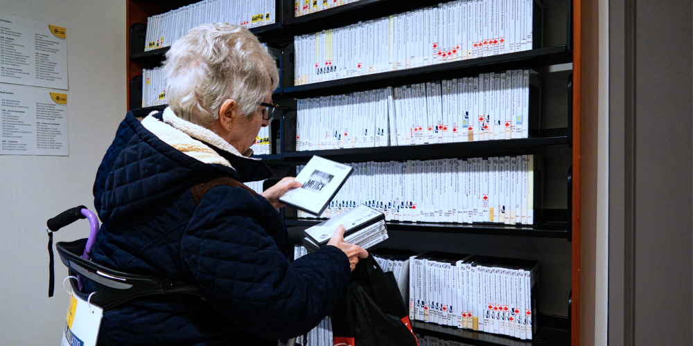 older adult using a rollator browsing a shelf full of DAISY disks, and holding six DAISY books in their hands.