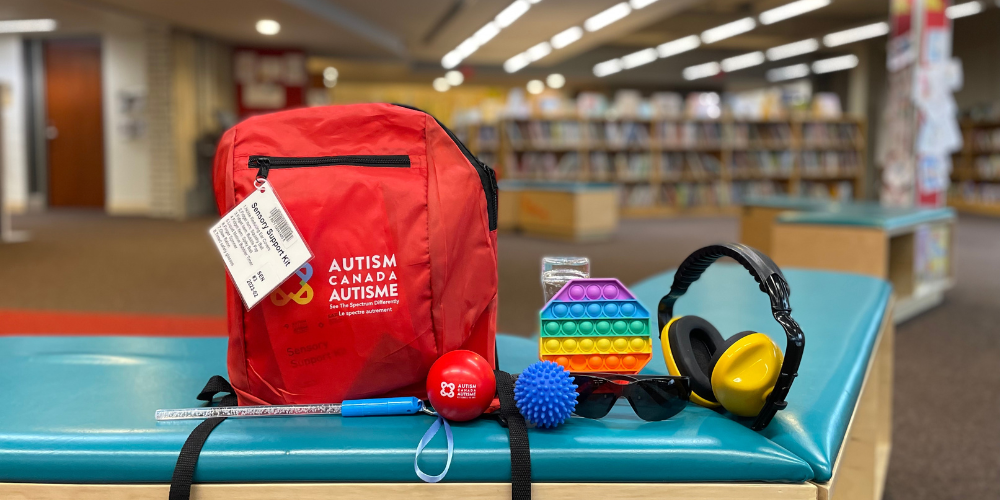 Red Autism Canada backpack resting on a green bench with two balls, headphones, a pop-it, and sunglasses resting beside it