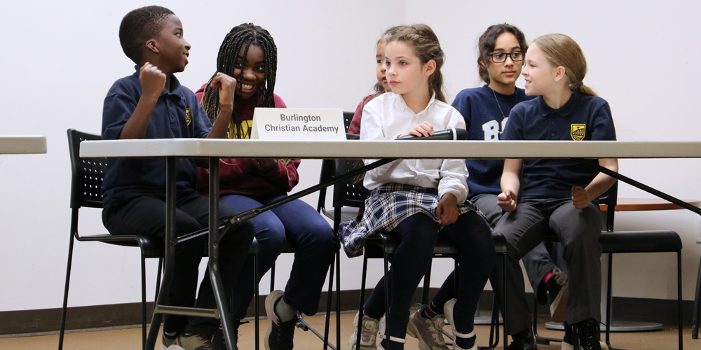 a team of six students sitting at a table talking excitedly