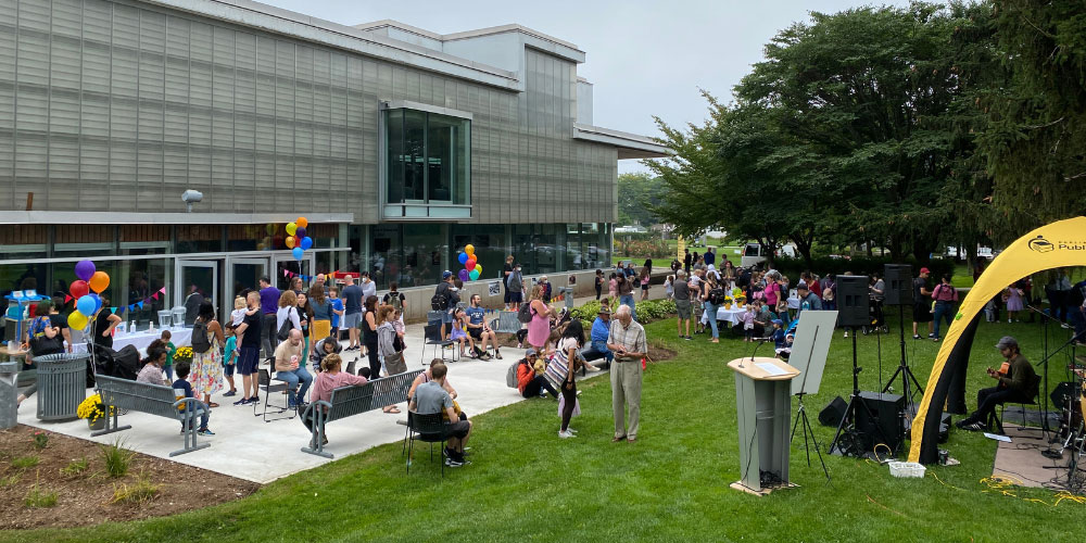 Exterior of library with a crowd of people gathered in front. Balloons and a yellow tent are in the image.