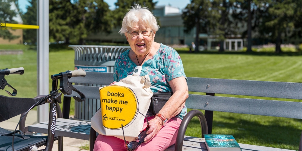 older adult sitting on a bus stop bench, beside their rollator, holding a library book bag