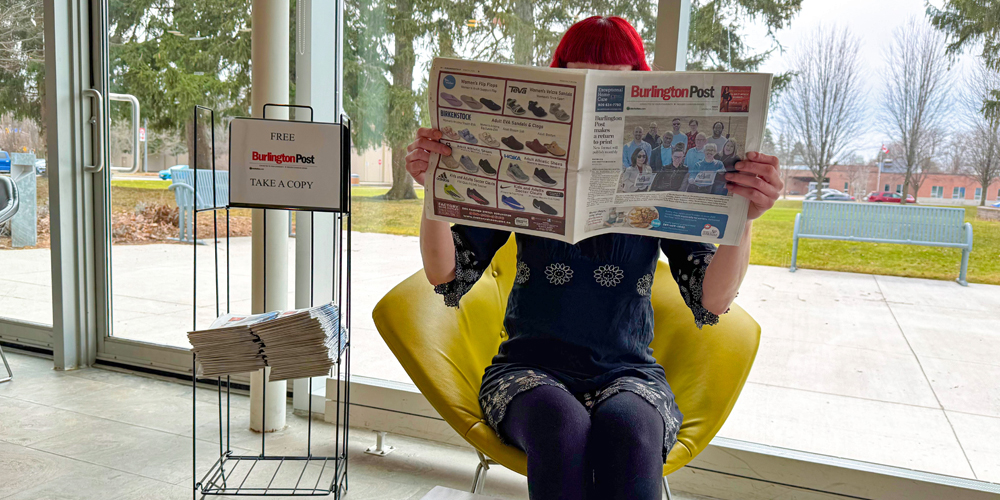 seated adult holding up a copy of The Burlington Post newspaper