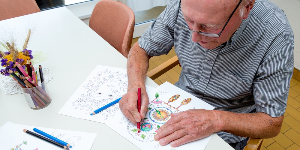 older adult sitting at a table using pencil crayons to colour in a design on a piece of paper