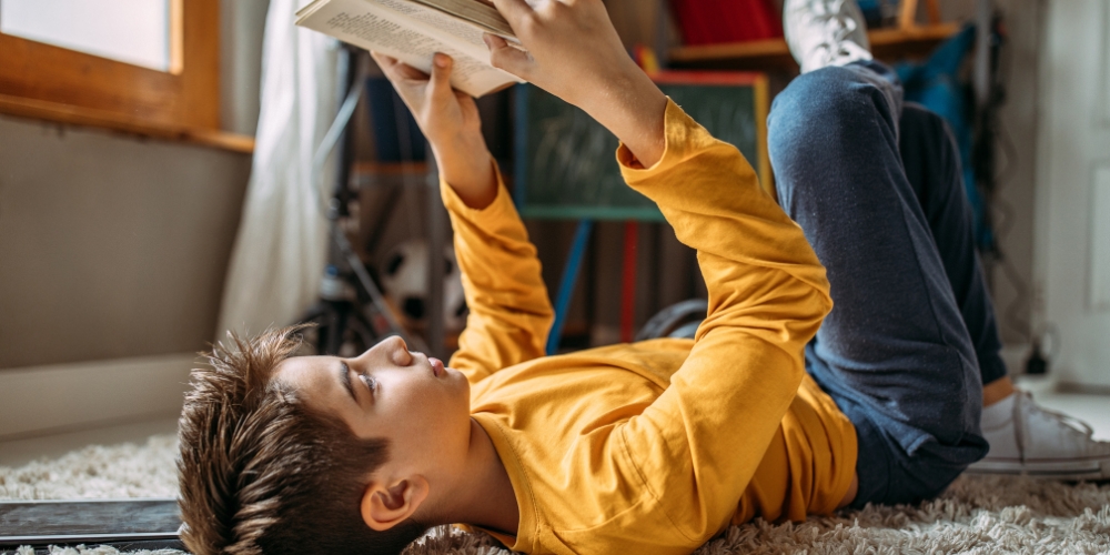 youth lying on floor beside a tablet and reading a book