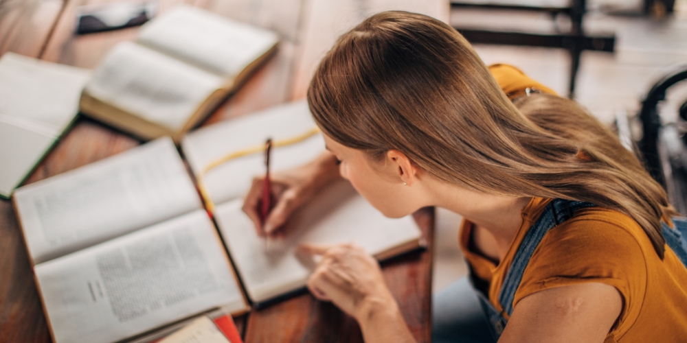 young adult sitting at a table writing in a notebook while looking at a textbook