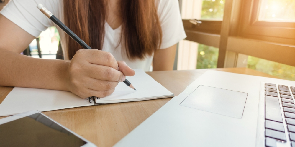 student sitting at a table with a laptop and tablet and writing on paper