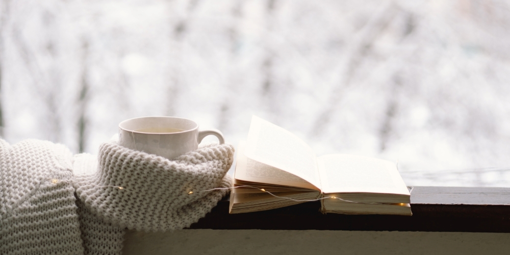 a tea cup wrapped in a knit scarf beside a book, all placed on a window ledge