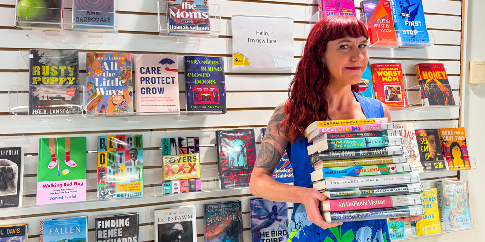 adult holding a stack of books and standing in front of a wall display of new books