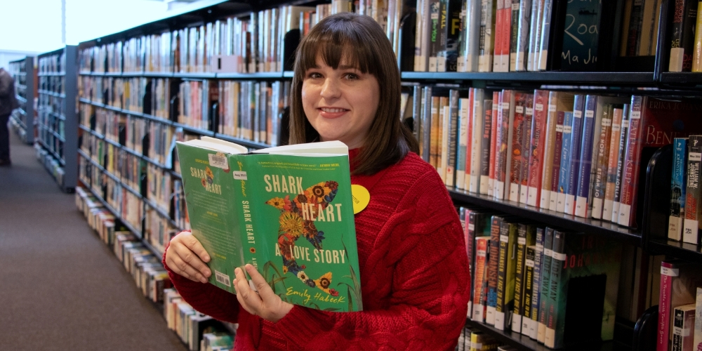 Librarian Emma standing in front of bookshelf holding book called Shark Heart, A Love Story by Emily Habeck 