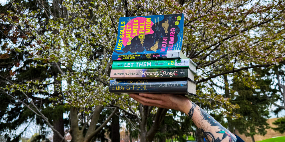 hand holding up four stacked books with a flowering tree in the background.