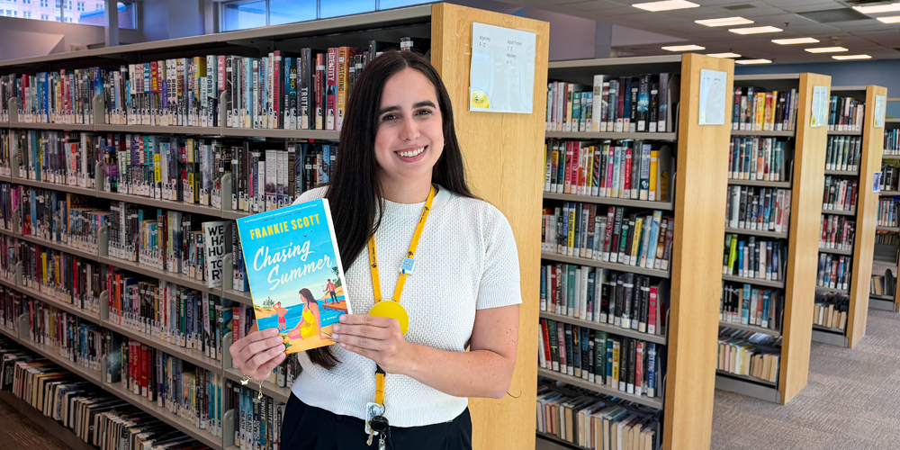 Librarian Astrid standing in front of book stacks and holding the book entitled Chasing Summer
