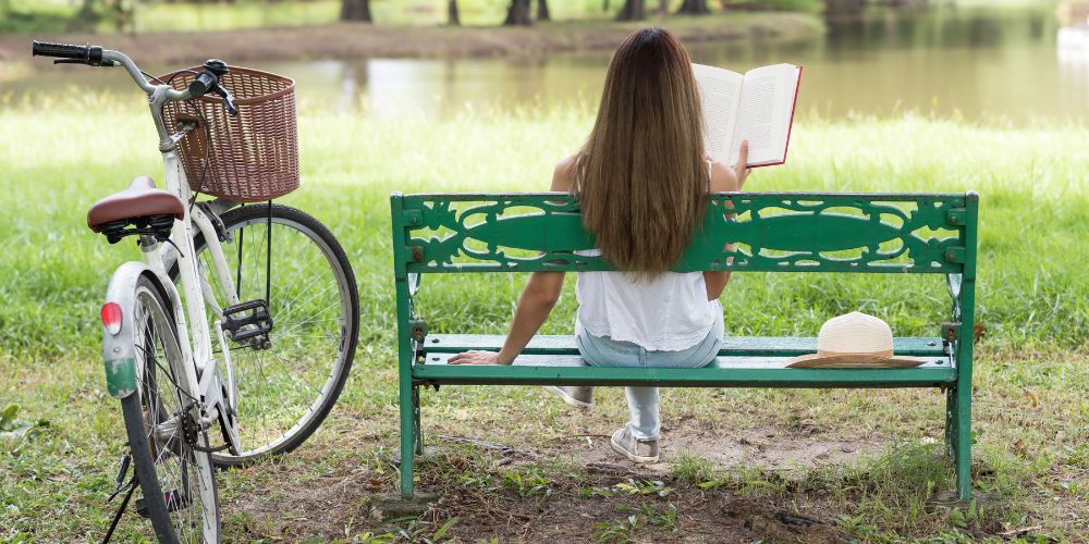 Woman sitting on a bench overlooking a pond while reading a book. Her bicycle is parked beside her.