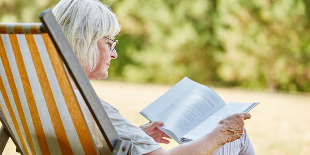 older adult sitting outside in a striped lawn chair and looking at a book