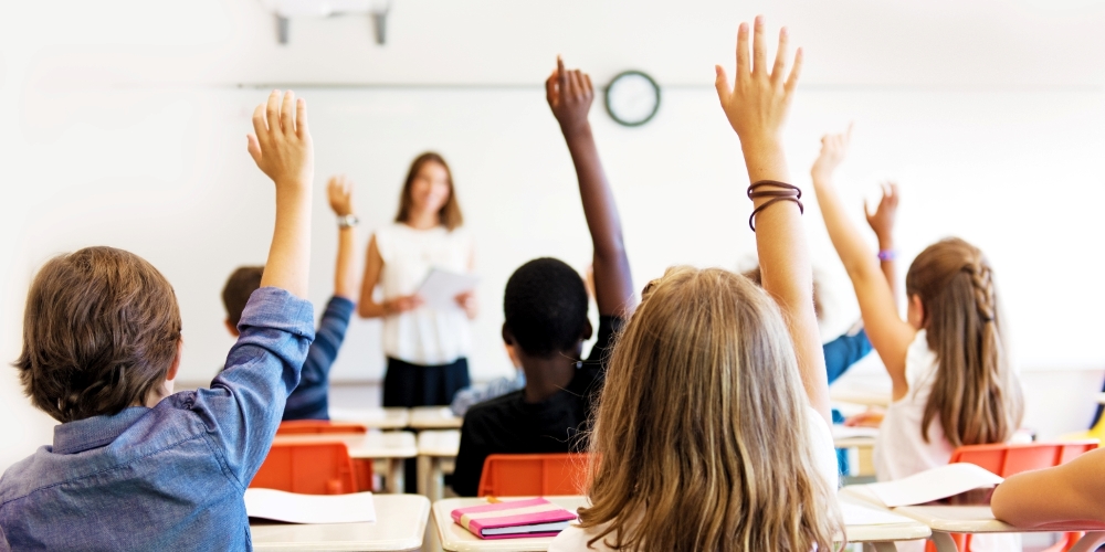 childen sitting in a classroom facing their teacher with their hands up.