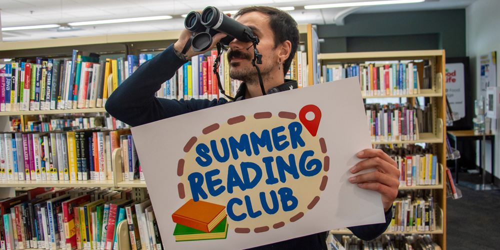 BPL staff looking through binoculars and holding a sign reading Summer Reading Club