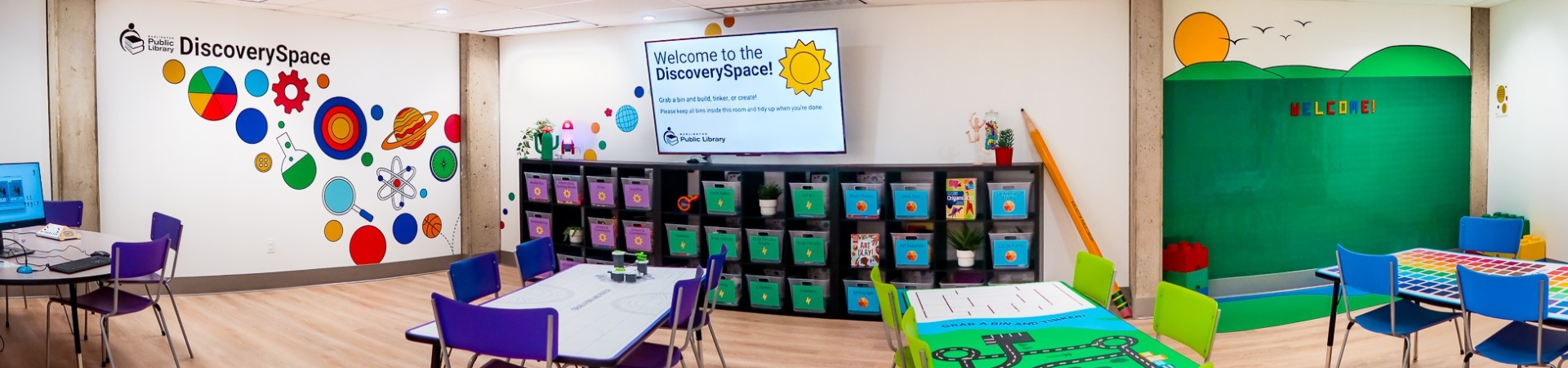 panorama of the DiscoverySpace showing four activity tables, self-directed activity bins, large tv screen, and LEGO wall
