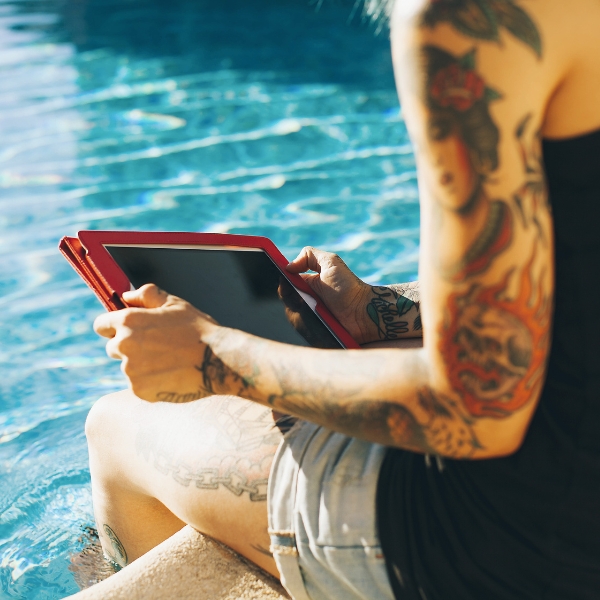 tattooed adult sitting poolside and holding a tablet 